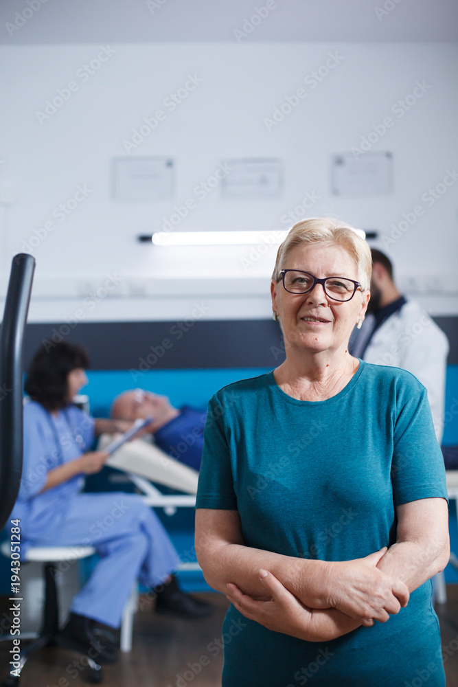 Fototapeta premium Old woman in portrait waits to work out for physical treatment with arms crossed. Senior patient with muscular injury gets ready for leg physiotherapy on stationary bike while she looks at camera.