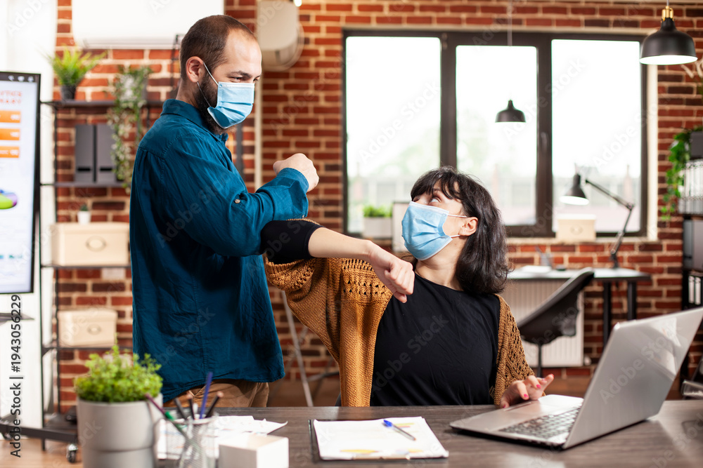 Fototapeta premium Businesswoman working at desk as male coworker greets her with elbow in brick wall office. Businessman and female colleague wearing face mask and maintaining safety in startup company.