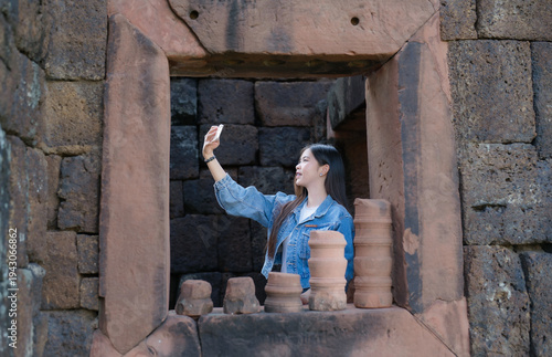 A young traveler stands inside an ancient stone doorway, taking a selfie with her smartphone while exploring a historic temple site. Surrounded by old stone walls and artifacts.