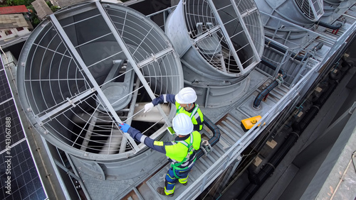 Two technicians in safety gear inspect large industrial cooling tower fans on a rooftop