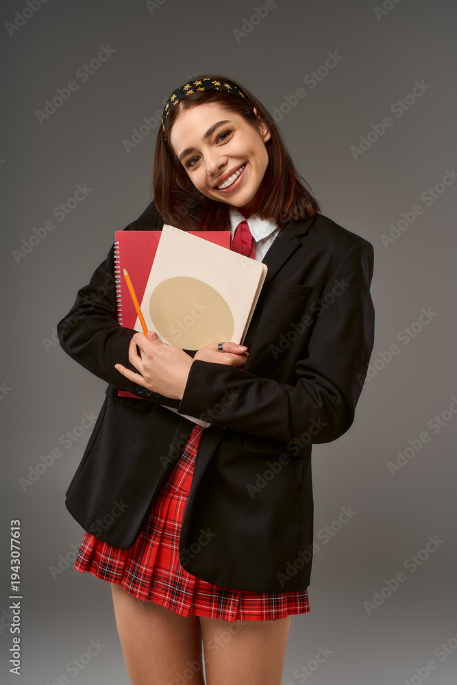 Fototapeta premium Joyful college girl in uniform holding books and smiling in studio setting