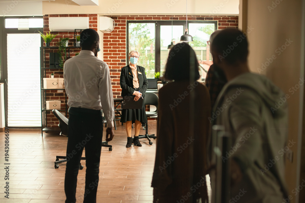 Fototapeta premium Elderly female therapist standing in brick wall room and welcoming clients inside, ready for therapy session. Group of people entering modern space as senior woman with face mask waits.