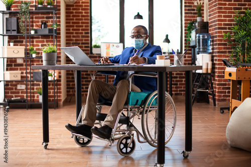 Wallpaper Mural Businessman in wheelchair using laptop and writing notes at office desk, preparing business project presentation. Black male employee with disability doing company research in brick wall workspace. Torontodigital.ca