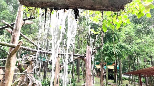Artificial waterfall in a lush green city park or zoo, featuring wooden structures and dence tropical trees in the background. Peaceful nature scene with flowing water.