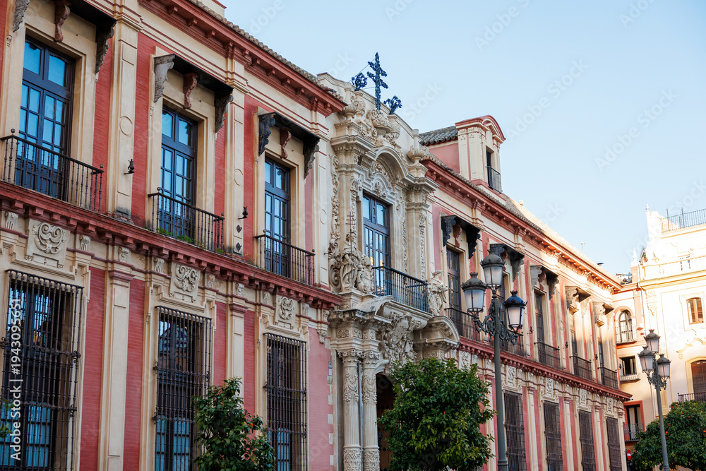 Obraz premium Archbishop's Palace Churrigueresque Portal, Seville, Spain. Street level view shows pink and cream pilasters, wrought iron balconies, carved entrance, cross, lampposts, and orange trees.