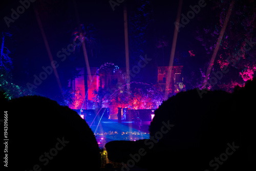 Seville nighttime light show in Moorish courtyard with palms, hedges, pond, fountains, and a small tower. Spotlights rise as jets sync, long exposure adds motion trails.