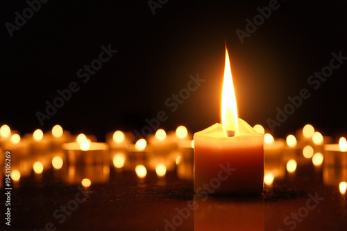 Burning candles as a symbol for religion or mourning in times of war. One large candleflame in front of defocused flames in the dark and black background.