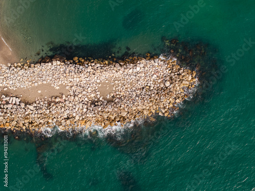 Fotografie Spain breakwater and sandy path seen aerial at midday, waves lap boulders, trian