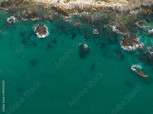 Aerial view of Spain coast shows turquoise water on rugged basalt rocks and a pale stone strip. Midday light reveals submerged boulders and swirling white foam.