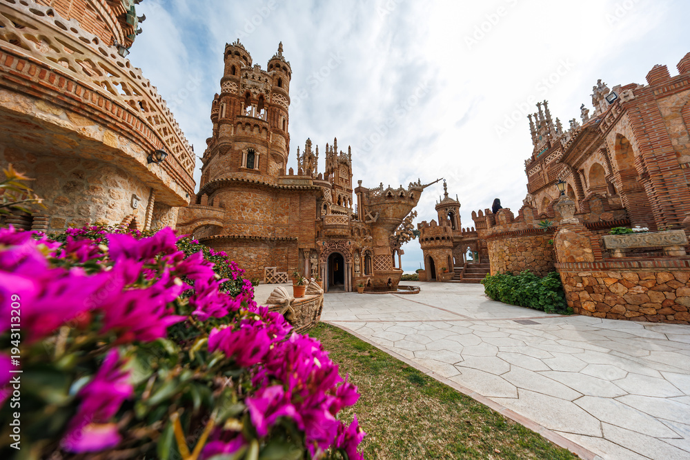 Obraz premium Ornate turrets and brickwork rise over a courtyard in Benalmadena, Malaga. Magenta bougainvillea, Mediterranean beyond arches, ship sculpture, Gothic and Mudejar motifs.