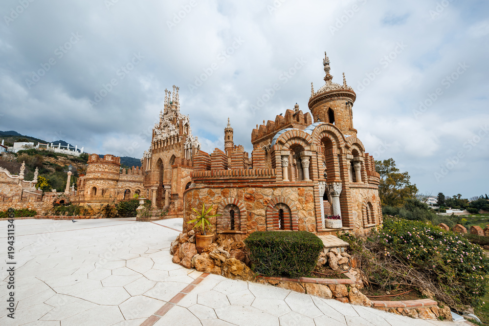 Obraz premium Castillo Monumento Colomares in Benalmadena, Malaga shows turrets, horseshoe arches, and ornate spires by a pale tiled courtyard under heavy clouds and even light.
