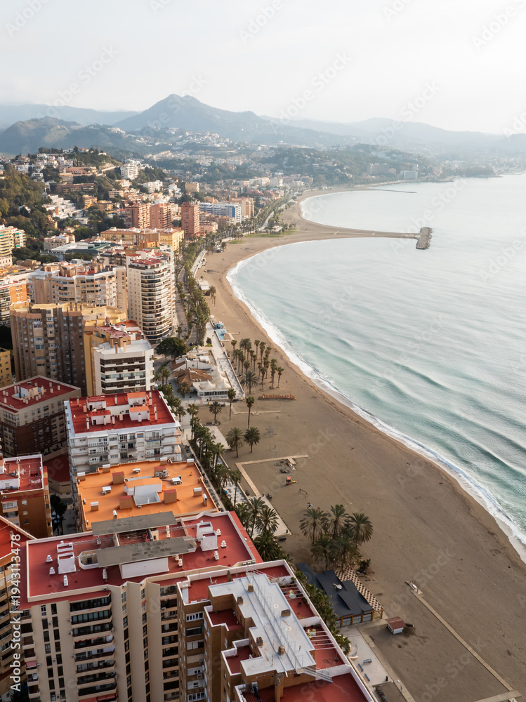 Fototapeta premium Aerial coastal view of Malaga, Spain shows La Malagueta and Pedregalejo, red roofed apartments, palm promenade, small breakwater, hazy Montes de Malaga, late afternoon light.