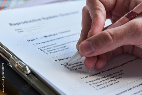 Close up of hand holding pen filling out reproductive system questionnaire on clipboard, marking answers on printed multiple choice test, focusing on education and assessment