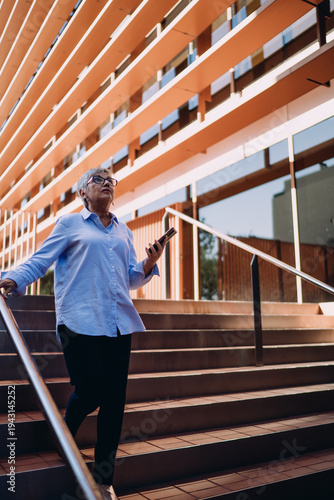 Schilderij op canvas Senior woman with short gray hair walking down steps holding smartphone, looking around with curiosity