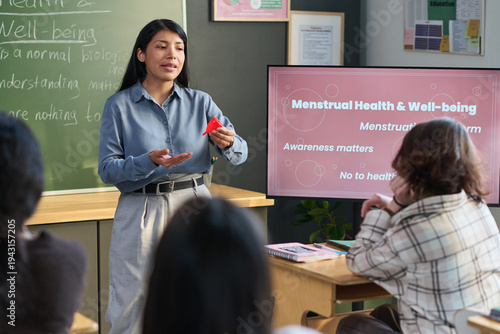 Young adult Hispanic woman standing in classroom holding menstrual cup, explaining menstrual health to group of teenagers seated at desks, educational presentation on screen