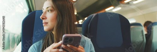 Cute girl with long brown hair, dressed in blue shirt, looks out window while traveling by suburban train, scrolls on smartphone screen. Panoramic