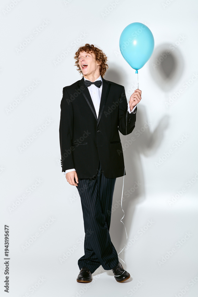 Obraz premium Curly-haired young man in a tuxedo celebrates with a blue balloon in a studio