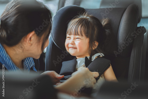 A loving Asian mother carefully buckles her smiling toddler daughter into a secure car seat before a road trip. Concept of child safety, parenting, and family travel security