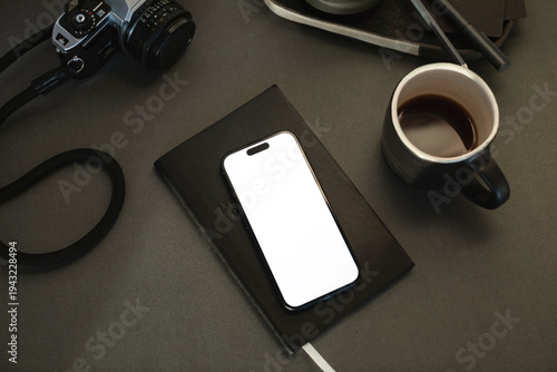 Close-up of a mobile phone with a blank screen on a notebook with a cup of black coffee and camera on a desk