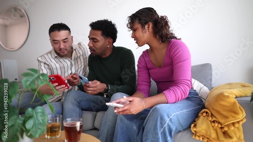 Three multi ethnic friends sitting on a sofa, happily watching and discussing something on their mobile phones while enjoying drinks in a cozy living room, fostering connection and shared moments