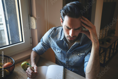 Concerned man feeling stressed and having headache while thinking and writing notes in a notebook, managing work or personal challenges at a home working desk