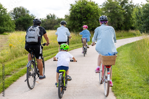 Family children cycling paved countryside path green meadow trees during warm summer day light. Active outdoor lifestyle parenting recreation freedom childhood adventure healthy travel concept