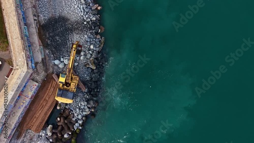 A yellow digger is near the water, moving rocks along the shoreline. Construction workers are present, and the scene shows a busy coastal area with clear water.
