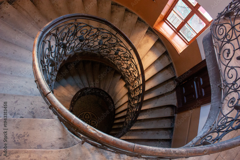 Fototapeta premium Elegant spiral staircase ornate wrought iron railing stone steps illuminated natural light window.