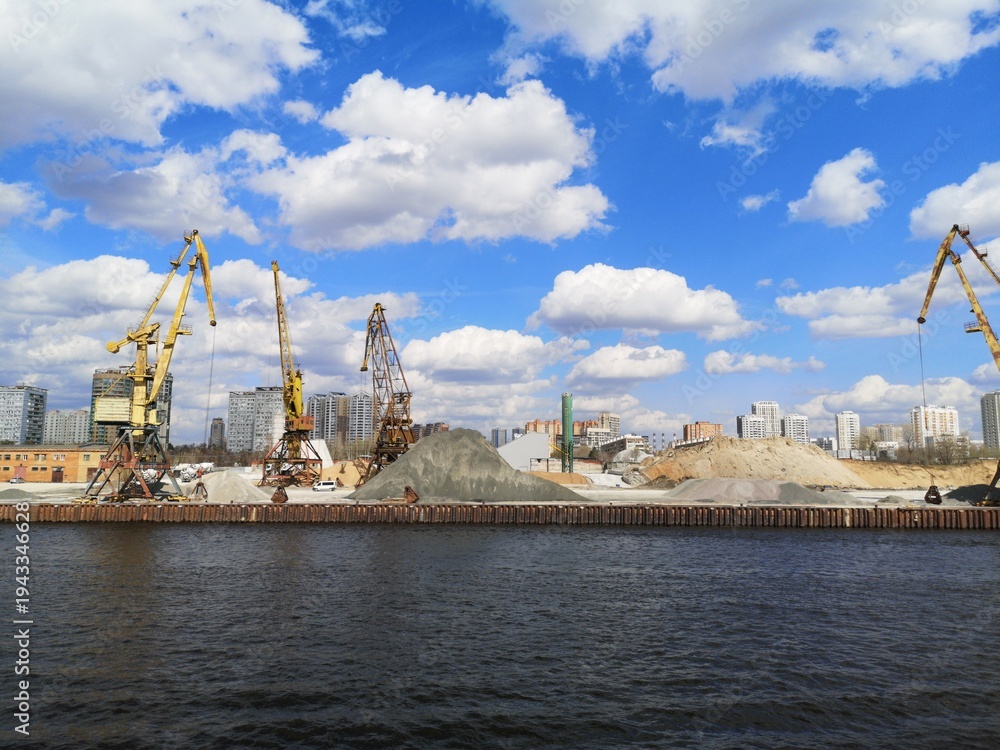Fototapeta premium Industrial river cargo port with cranes, sand piles and city skyline under blue sky with clouds.