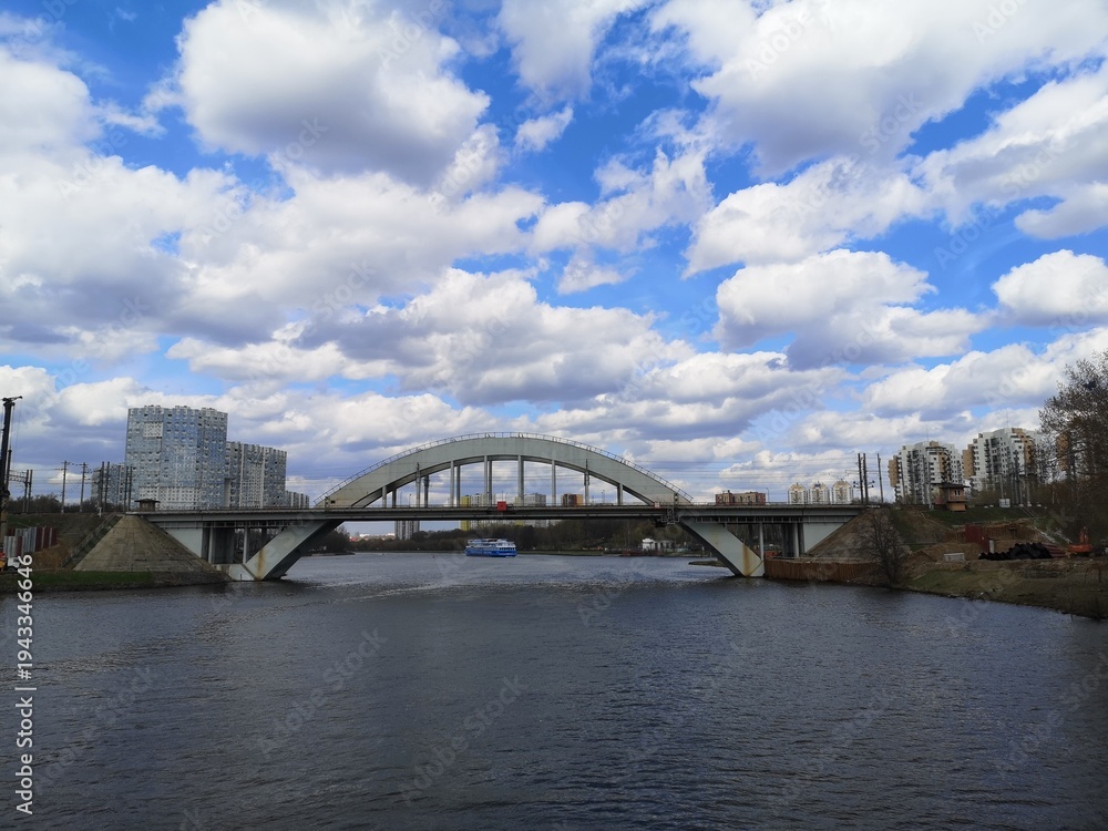 Fototapeta premium Urban arched bridge spanning a calm river with modern buildings nearby and dramatic clouds in a blue sky cityscape.