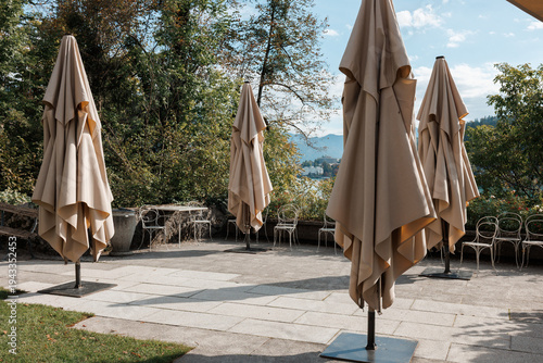 Canvas Print Large beige sun umbrellas remain closed on an empty stone cafe terrace overlooking the trees and water of Lake