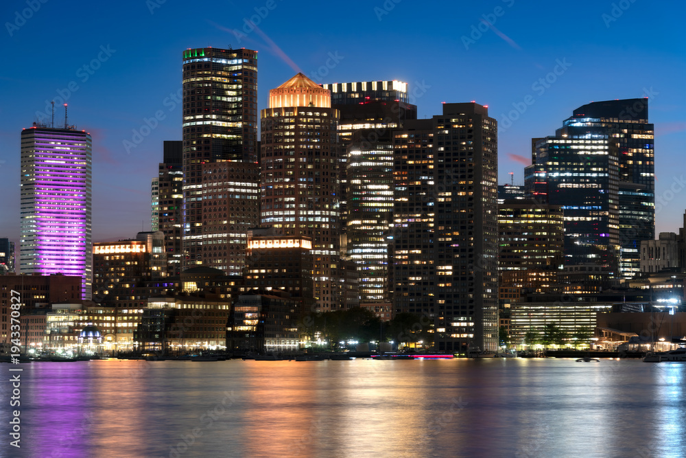 Obraz premium The illuminated skyscrapers of downtown Boston reflect in the calm waters of the harbor during the blue hour.