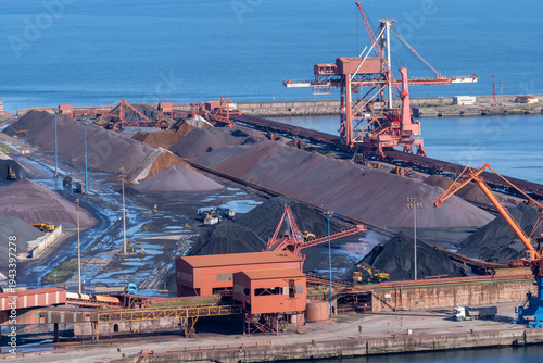 A wide view of the mineral storage area at the Port of Gijon, Asturias, featuring large piles of coal and iron ore with industrial conveyors and port cranes.