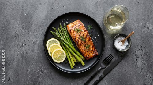 Top-down view of grilled salmon, asparagus, and lemon on a black plate, served with white wine and sea salt on a dark grey concrete background.