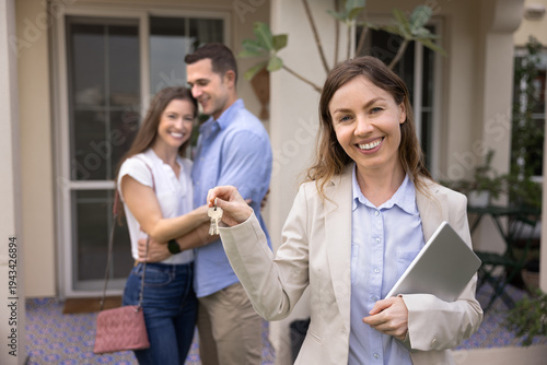 Foto Portrait of smiling female realtor holding house keys, symbolizing completion of successful home purchase, happy couple stands in background