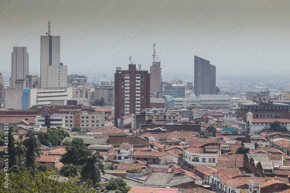 Obraz premium A panoramic view of Cali, Colombia, contrasting the red-tiled roofs of the historic district with the towering skyscrapers of the modern city center under a hazy, overcast sky.