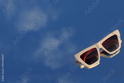 Bilde på lerret Stylish white cat-eye sunglasses reflecting the clear blue sky on a mirrored sur