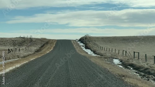 First person perspective driving on isolated prairie gravel road beneath expansive blue sky in rural Alberta Canada.