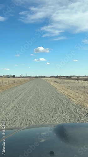 Point of view driving on rural gravel road through open prairie landscape under blue sky in Alberta Canada.