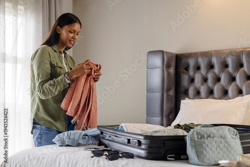 Mid-adult Indian woman standing beside bed in hotel bedroom, holding blouse and packing suitcase