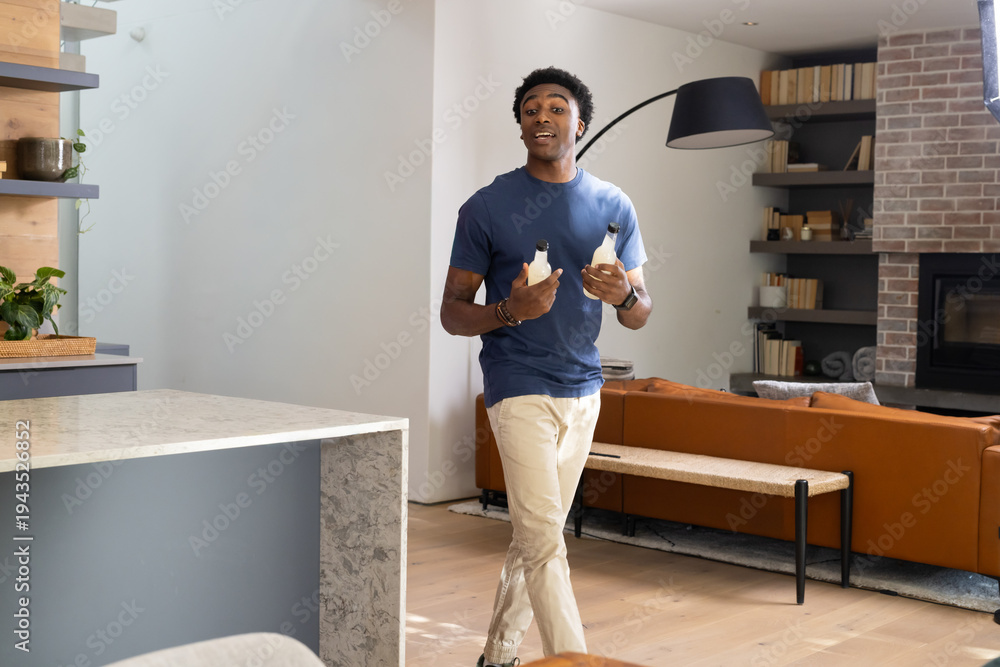 Fototapeta premium African American man walking through open-plan home holding two beverage bottles near marble island