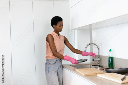 African American woman standing on left washing frying pan at kitchen sink wearing pink gloves