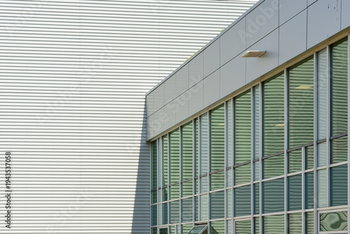The corner of a large grey building with metal composite panels and a row of commercial glass windows. There's a larger white exterior metal-clad siding wall of a building in the background.