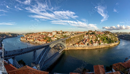 A sweeping cityscape view of a river, bridge, and clustered buildings under a clear, cloud-streaked blue sky