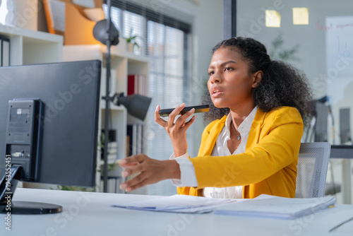 Confident professional businesswoman using smartphone voice message while reviewing documents at modern office desk. Young corporate employee working with technology, communication tools, and producti