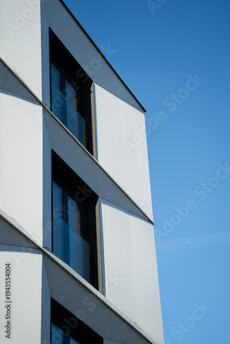 Geometric white facade of a modern building with large windows and sharp lines, set against a clear blue sky. Suitable for illustrating contemporary architecture, urban design, or real estate concepts