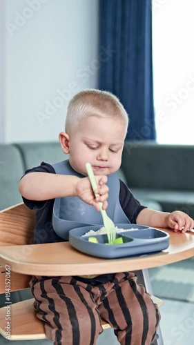 Adorable 2 year old boy sits at home and eats independently. Toddler learns to eat independently. Child in a high chair sits and eats porridge deliciously.
