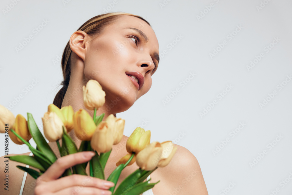 Fototapeta premium A young woman with clear skin holds a bouquet of yellow tulips close to her chest while looking thoughtfully to the side against a plain light background, beauty concept.