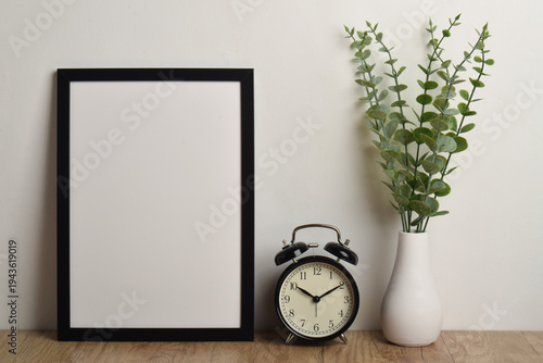 Photo frame mockup, alarm clock and plant vase on wooden table.