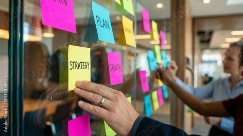 Dynamic close-up of business professionals actively collaborating, placing vibrant sticky notes with strategic plans onto a transparent glass board in a modern office